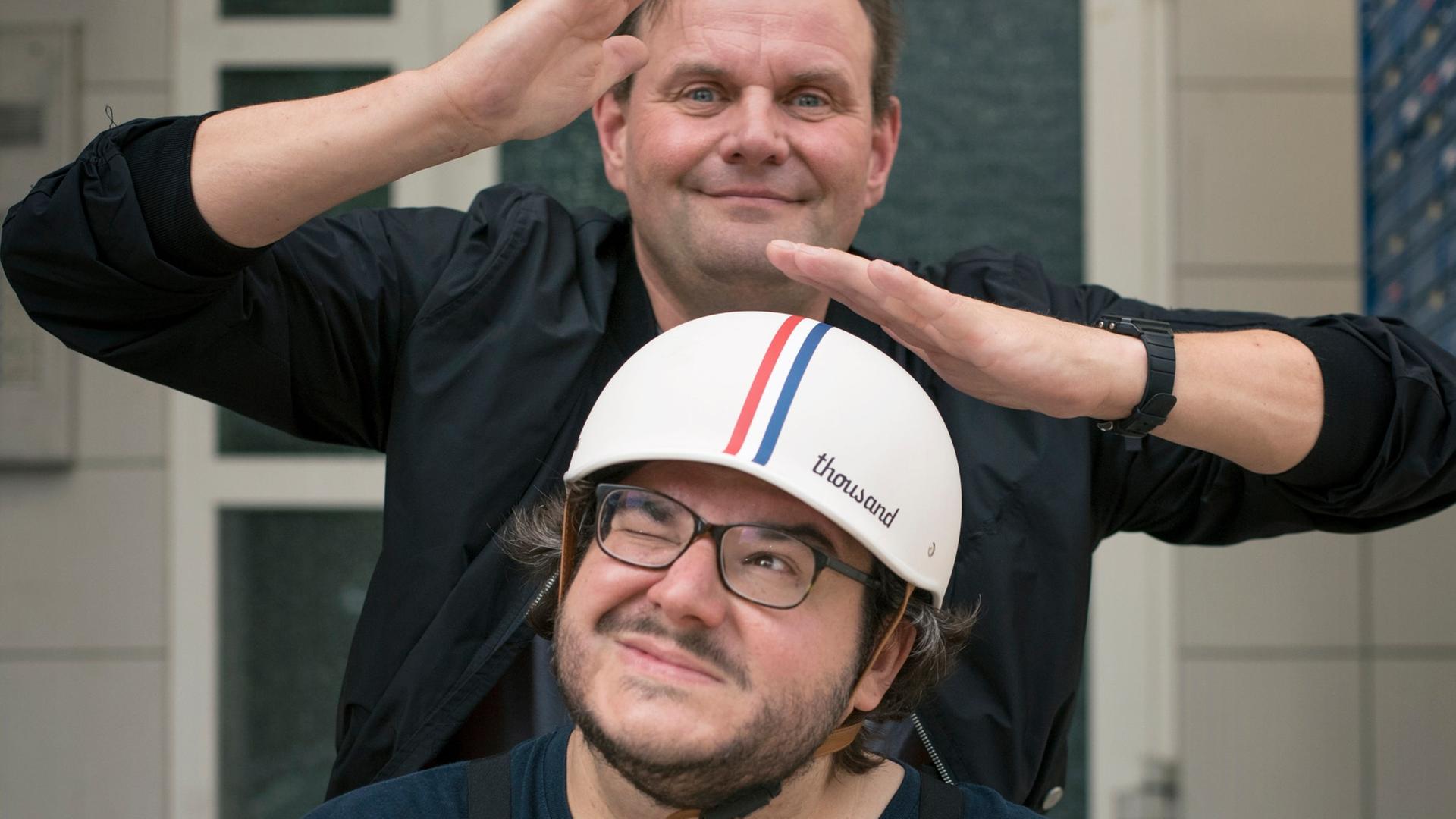 Devid Striesow und Axel Ranisch stehen hintereinander, Axel Ranisch trägt einen Mopedhelm und schaut zur Seite. Devid Striesow hält die Hände über seinen Kopf und schaut albern in die Kamera. Devid Striesow und Axel Ranisch stehen hintereinander, Axel Ranisch trägt einen Mopedhelm und schaut zur Seite. Devid Striesow hält die Hände über seinen Kopf und schaut albern in die Kamera.