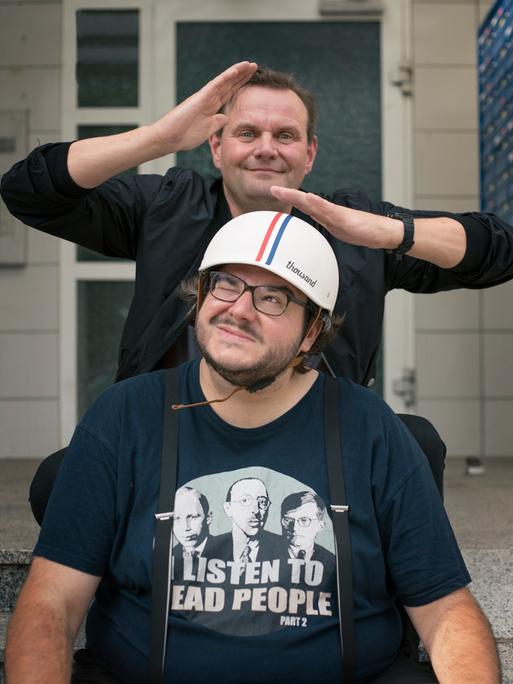 Devid Striesow und Axel Ranisch stehen hintereinander, Axel Ranisch trägt einen Mopedhelm und schaut zur Seite. Devid Striesow hält die Hände über seinen Kopf und schaut albern in die Kamera.