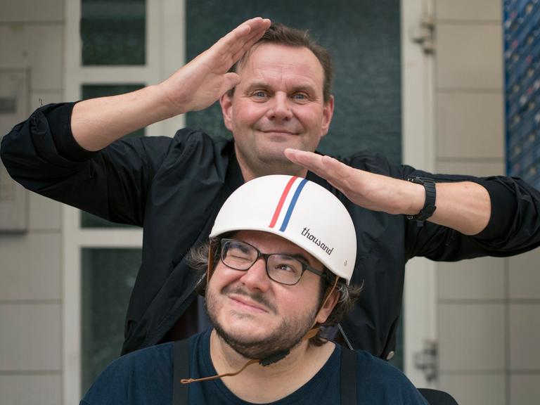 Devid Striesow und Axel Ranisch stehen hintereinander, Axel Ranisch trägt einen Mopedhelm und schaut zur Seite. Devid Striesow hält die Hände über seinen Kopf und schaut albern in die Kamera.