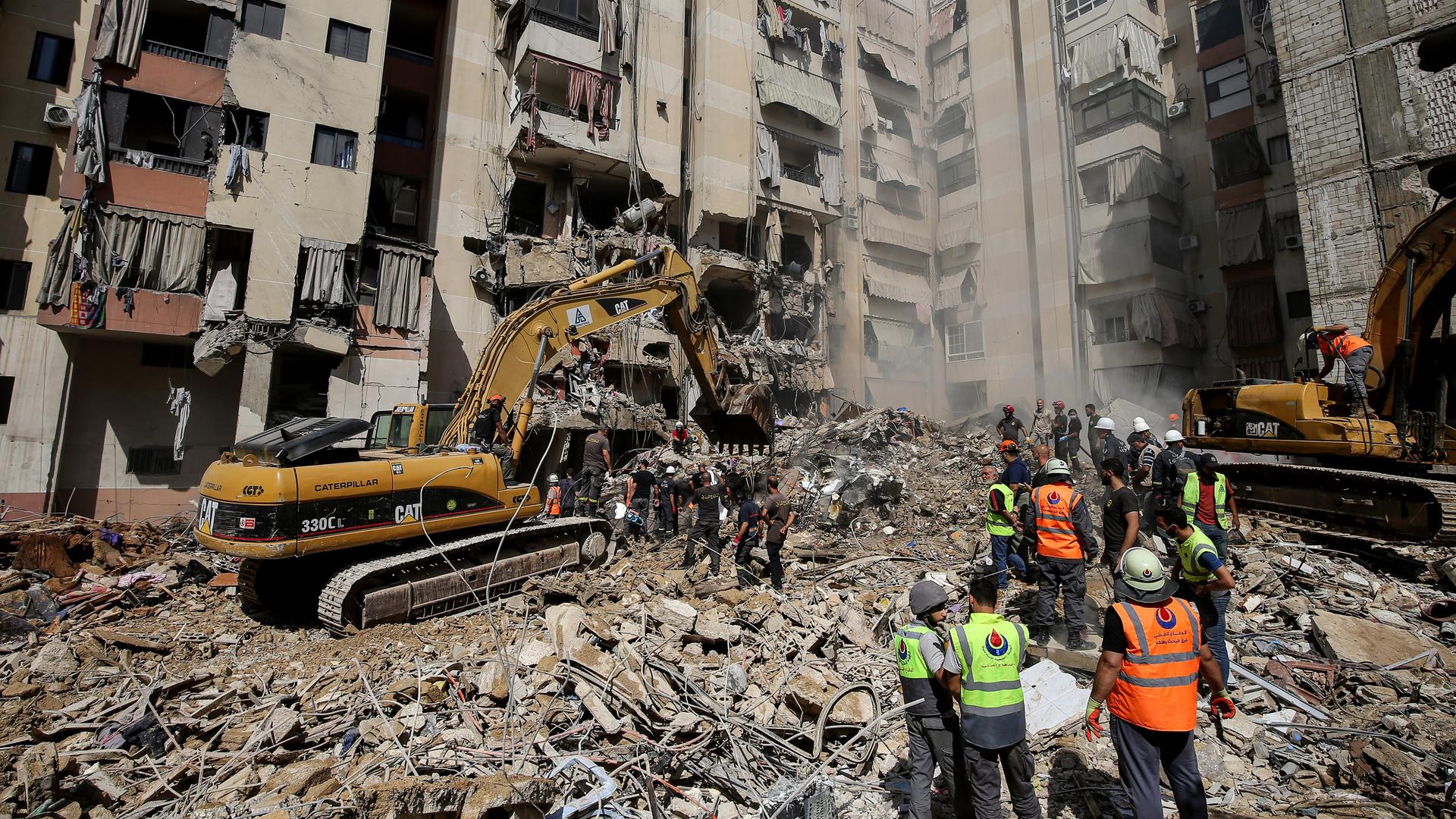 Männer in Schutzwesten und ein Bagger stehen auf einem Schuttberg inmitten einem Wohngebiet in Beirut. 