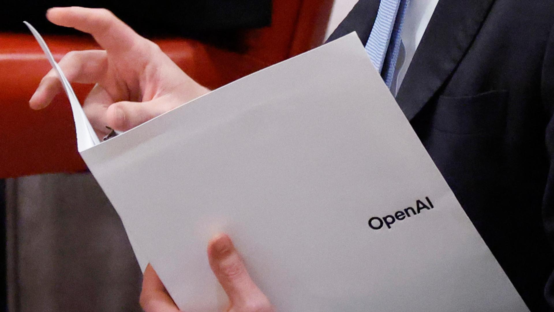 Ein Mann mit einem Anzug und einer lilafarbenen Krawatte, von dem man nur die Körpermitte sieht, hält einen weißen Papierordner in der Hand, auf dem "OpenAI" steht. Ein Mann mit einem Anzug und einer lilafarbenen Krawatte, von dem man nur die Körpermitte sieht, hält einen weißen Papierordner in der Hand, auf dem "OpenAI" steht.