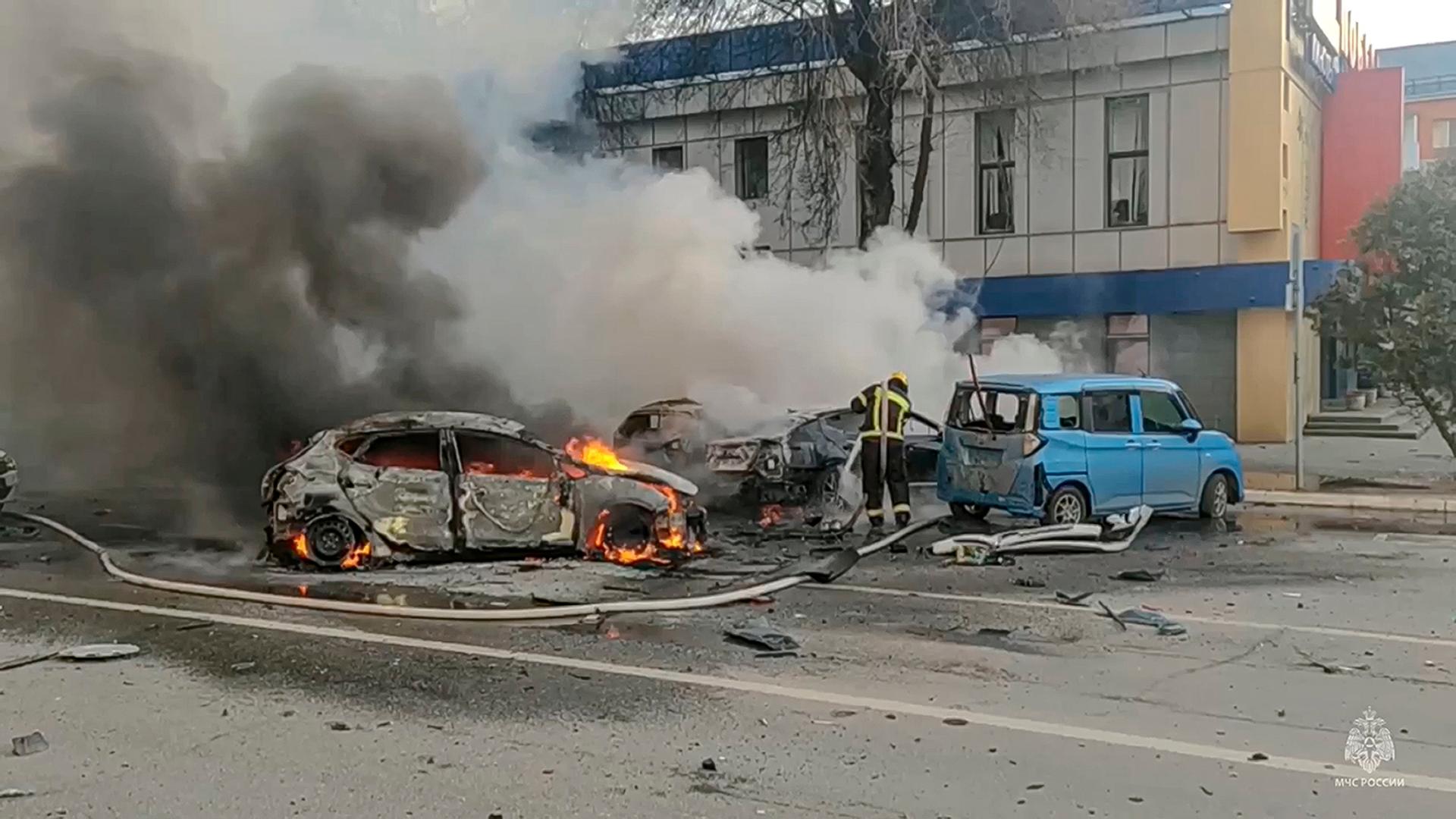 Feuerwehrleute löschen auf einer Straße zwei brennende Autos, aus denen dichter Qualm dringt. Dahinter ein mehrstöckiges Haus.