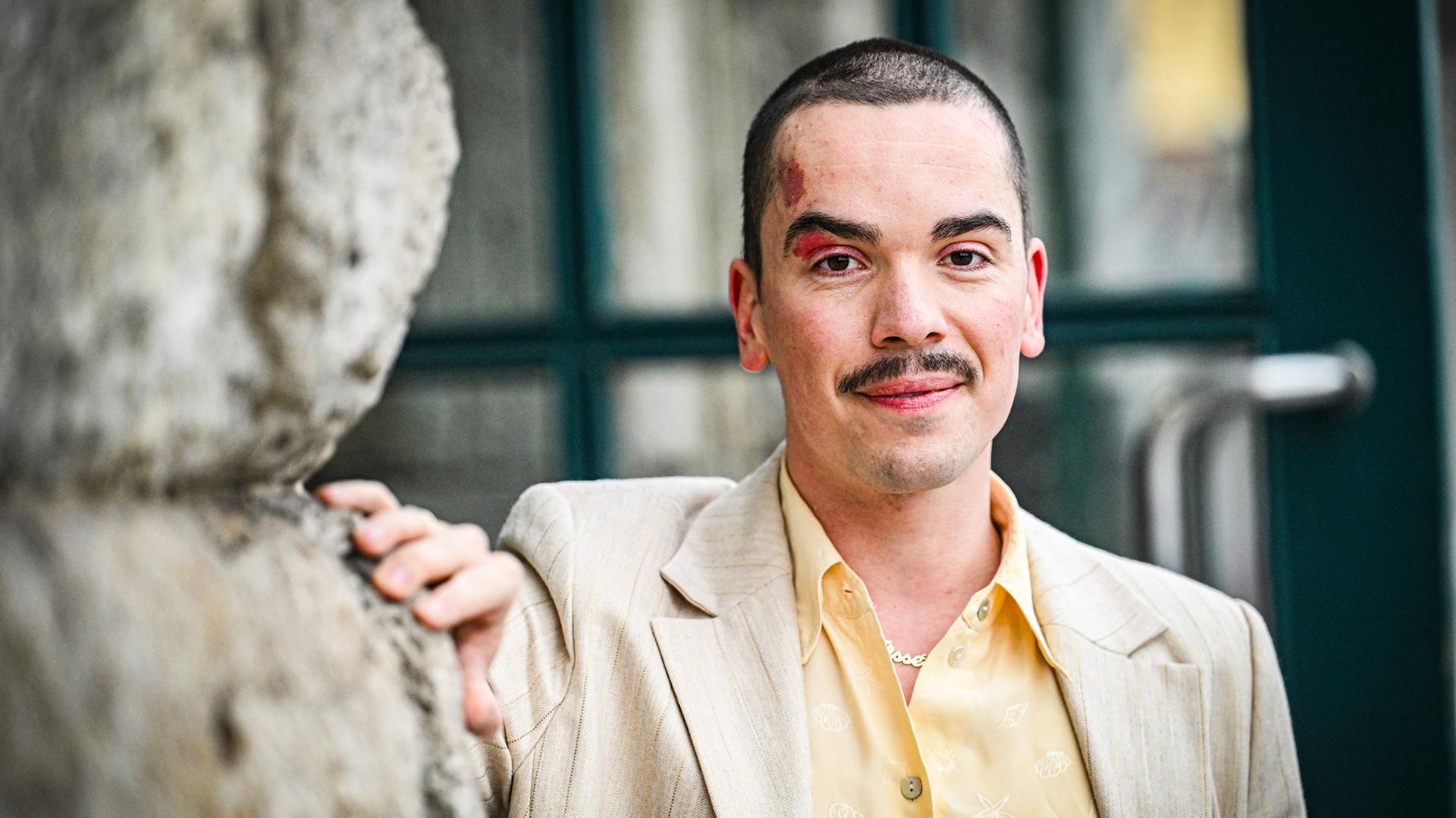 Ole Liebl steht an eine Wand gelehnt und schaut freundlich in die Kamera. Er trägt ein gelbes Hemd und helles Sakko.