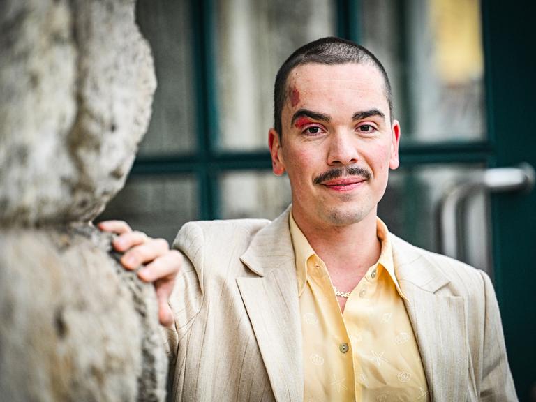 Ole Liebl steht an eine Wand gelehnt und schaut freundlich in die Kamera. Er trägt ein gelbes Hemd und helles Sakko.