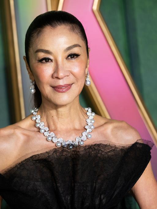 Michelle Yeoh lächelt und trägt ein schwarzes Kleid mit Perlen-Schmuck an Hals und Ohren. 