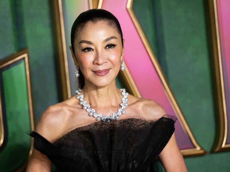 Michelle Yeoh lächelt und trägt ein schwarzes Kleid mit Perlen-Schmuck an Hals und Ohren. 