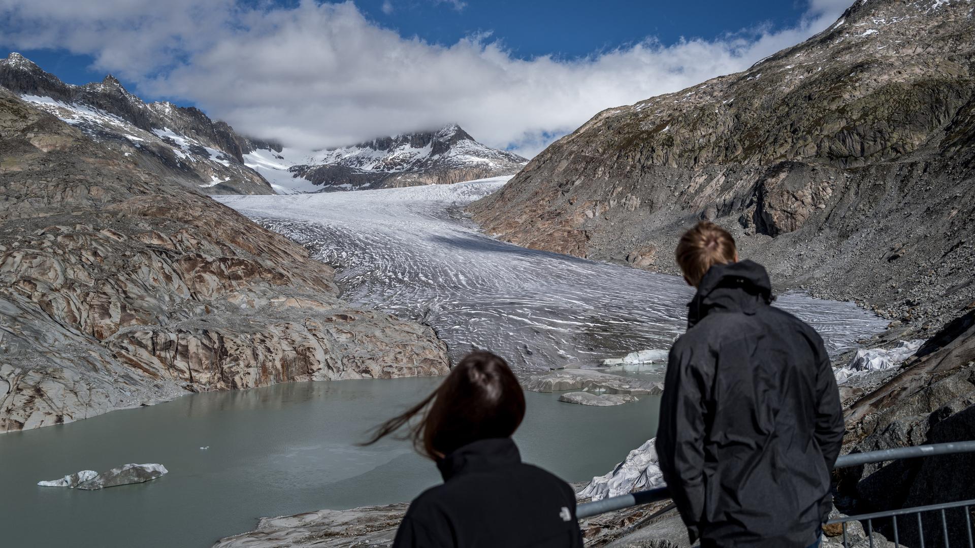 Zwei Touristen betrachten den Rhone-Gletscher, der sich zwischen zwei Berggipfeln hindurch schlängelt und in einem See endet.