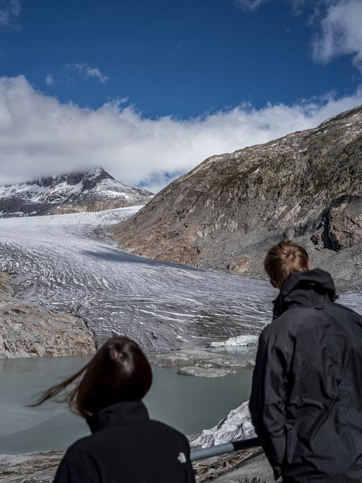 Zwei Touristen betrachten den Rhone-Gletscher, der sich zwischen zwei Berggipfeln hindurch schlängelt und in einem See endet.