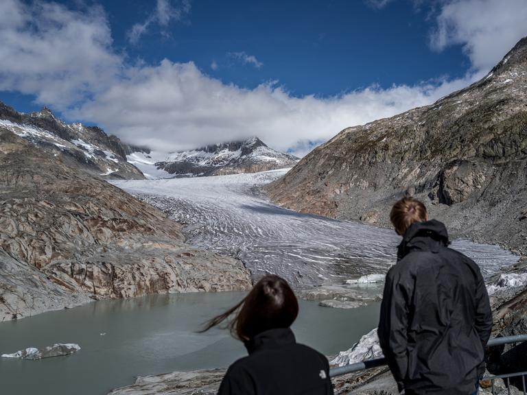 Zwei Touristen betrachten den Rhone-Gletscher, der sich zwischen zwei Berggipfeln hindurch schlängelt und in einem See endet.