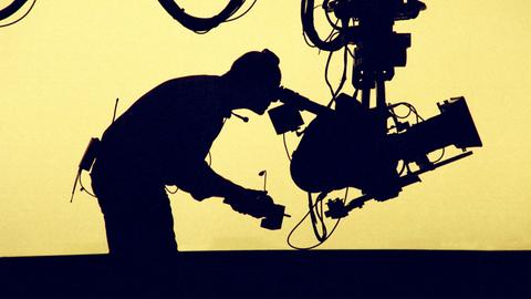 Ein Kameramann, der eine Kamera bedient. Er ist vor gelbem Gegenlicht nur als Silhouette erkennbar. Ein Kameramann, der eine Kamera bedient. Er ist vor gelbem Gegenlicht nur als Silhouette erkennbar.