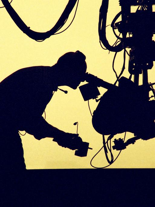 Ein Kameramann, der eine Kamera bedient. Er ist vor gelbem Gegenlicht nur als Silhouette erkennbar. Ein Kameramann, der eine Kamera bedient. Er ist vor gelbem Gegenlicht nur als Silhouette erkennbar.