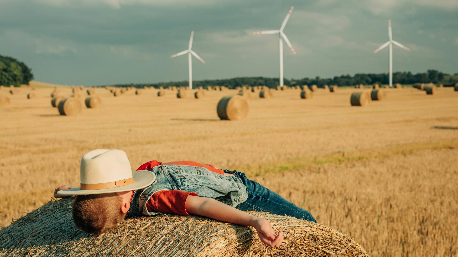 Ein Junge in Cowboy Kledung mit Hut und Jeans, liegend auf einem Heuballen in einem gemähten Weizenfeld, Symbolfoto.