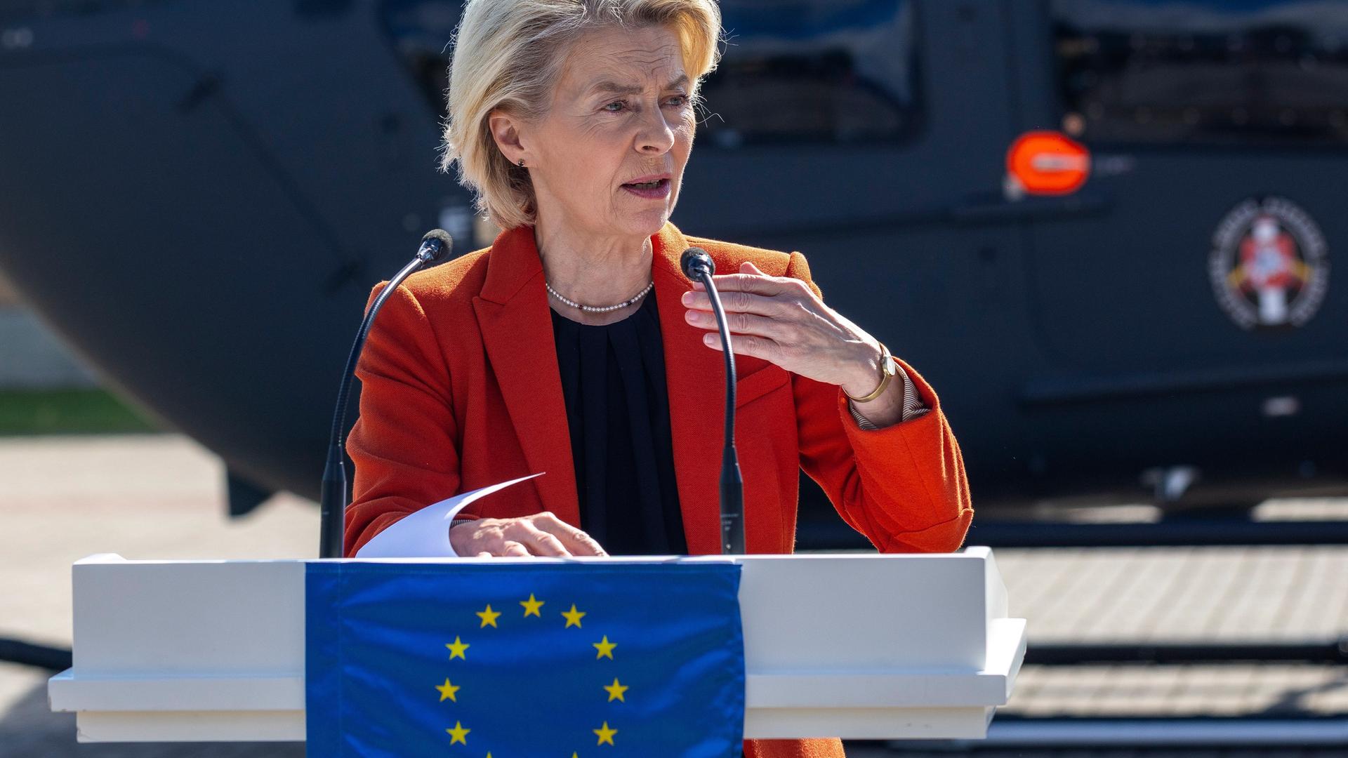 Die Präsidentin der Europäischen Kommission, Ursula von der Leyen, steht vor einem Rednerpult, das eine europäische Flagge ziert. Im Hintergrund steht ein Hubschrauber.