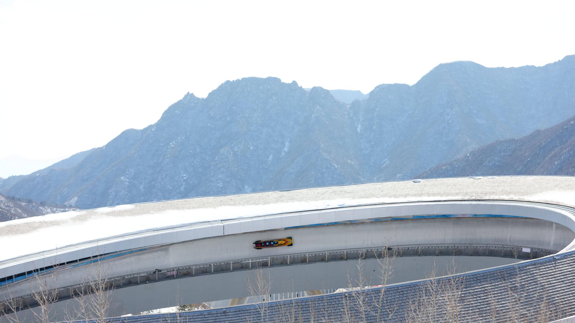 Ein Bobschlitten in einer Kurve auf der Bahn bei Peking, im Hintergrund sind Berge zu erkennen.