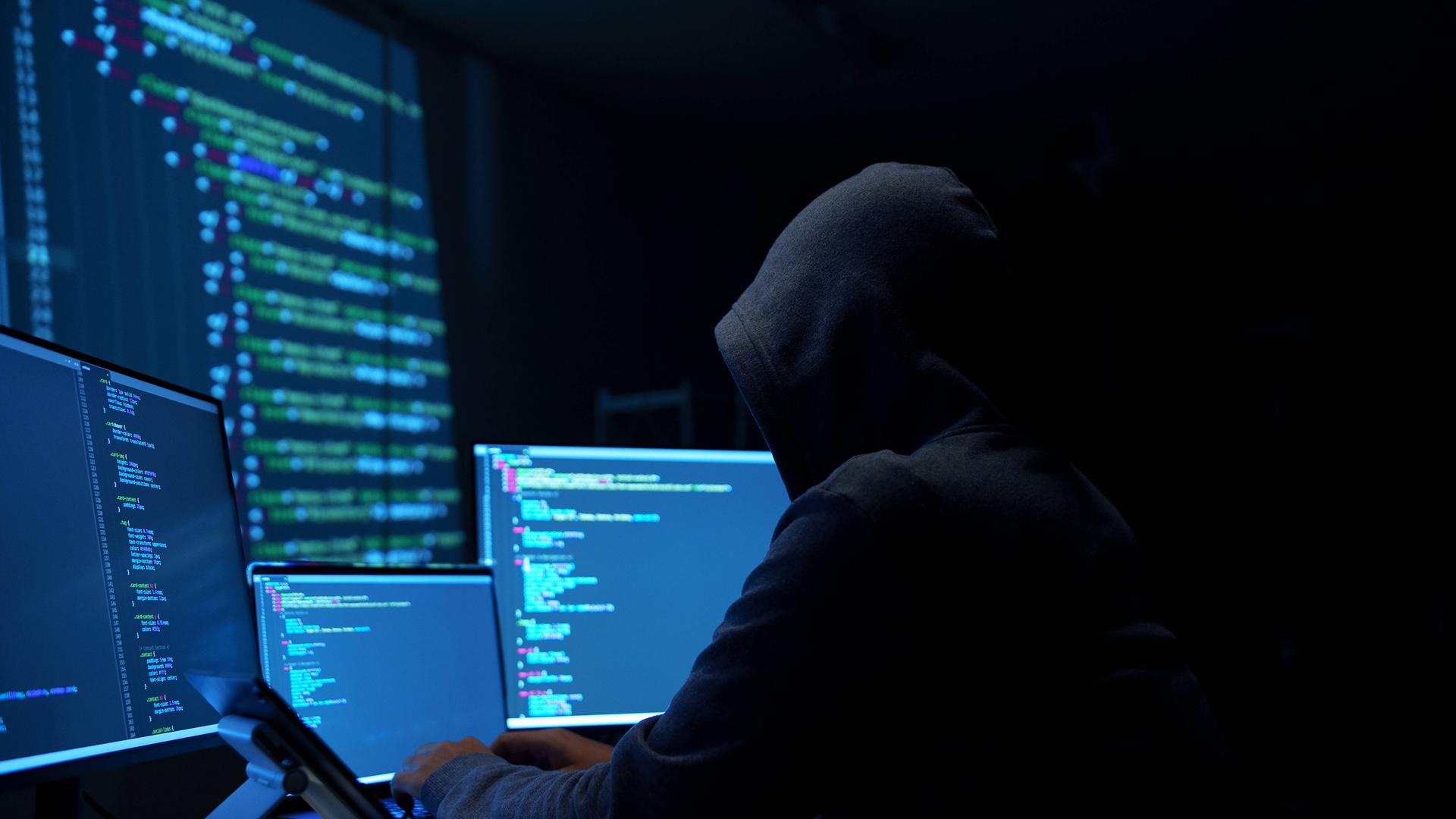 Ein Mann mit einem Kapuzen-Pulli sitzt vor mehreren Computer-Bildschirmen. Sein Gesicht ist nicht zu sehen.