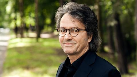 Ralf Rothmann schaut in die Kamera. Er hat dunkles Haar mit grauen Strähnen, trägt eine Brille und hat ein dunkles Hemd an. Im Hintergrund ist eine Parklandschaft zu erkennen.