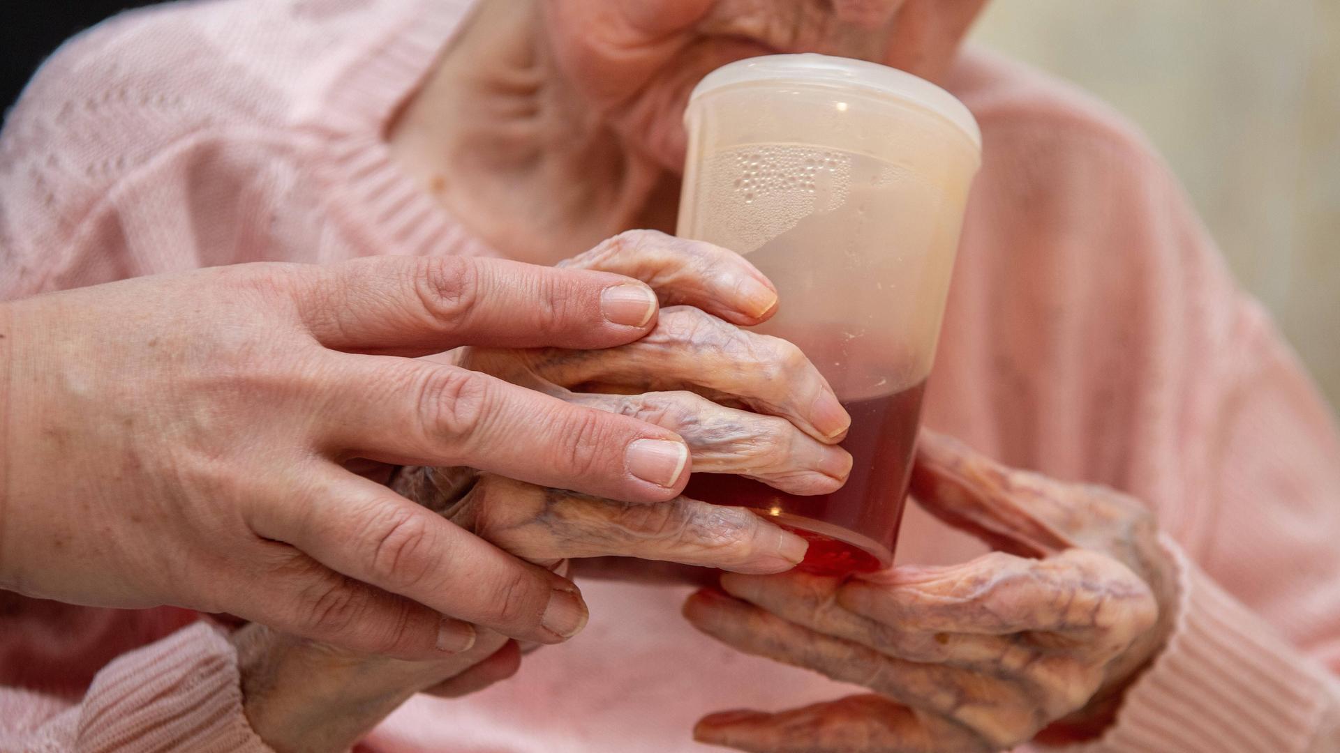 Eine Altenpflegerin hilft einer Seniorin beim halten eines Trinkbechers