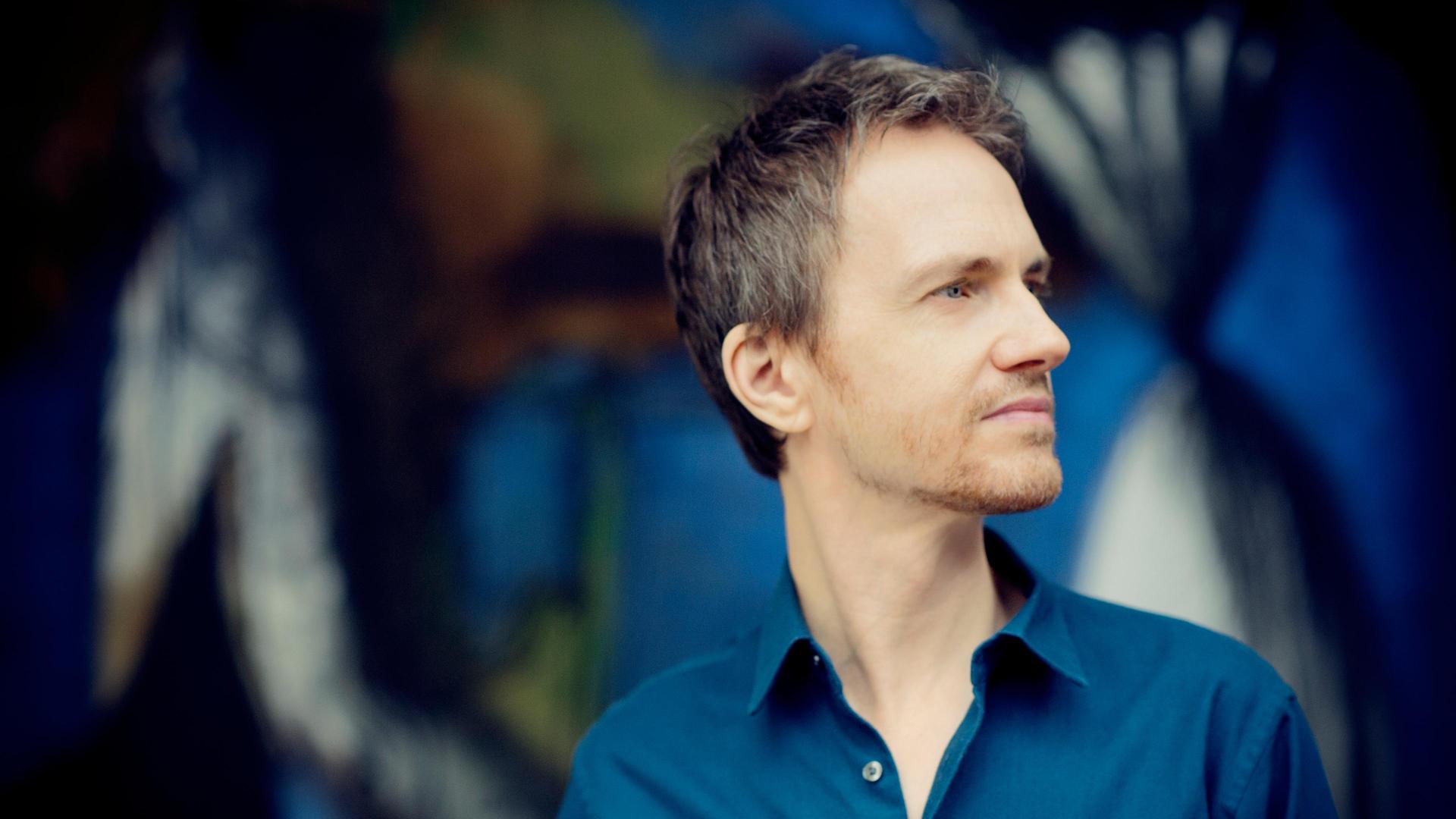 Alexandre Tharaud steht vor einer bunten Wand und schaut seitwärts in die Ferne.