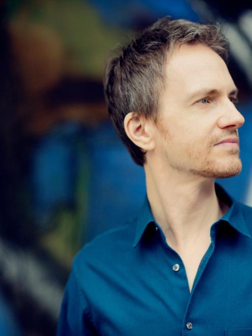 Alexandre Tharaud steht vor einer bunten Wand und schaut seitwärts in die Ferne.