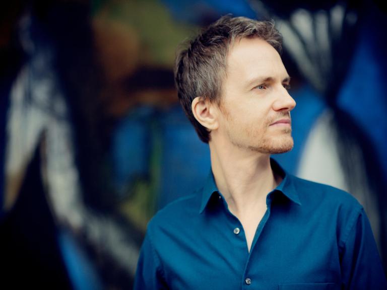 Alexandre Tharaud steht vor einer bunten Wand und schaut seitwärts in die Ferne.