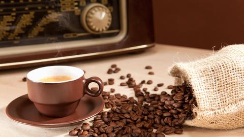 Eine Tasse Kaffee steht vor einem Radiogerät und neben einem umgefallenen Jute-Sack, aus dem Kaffeebohnen gefallen sein.