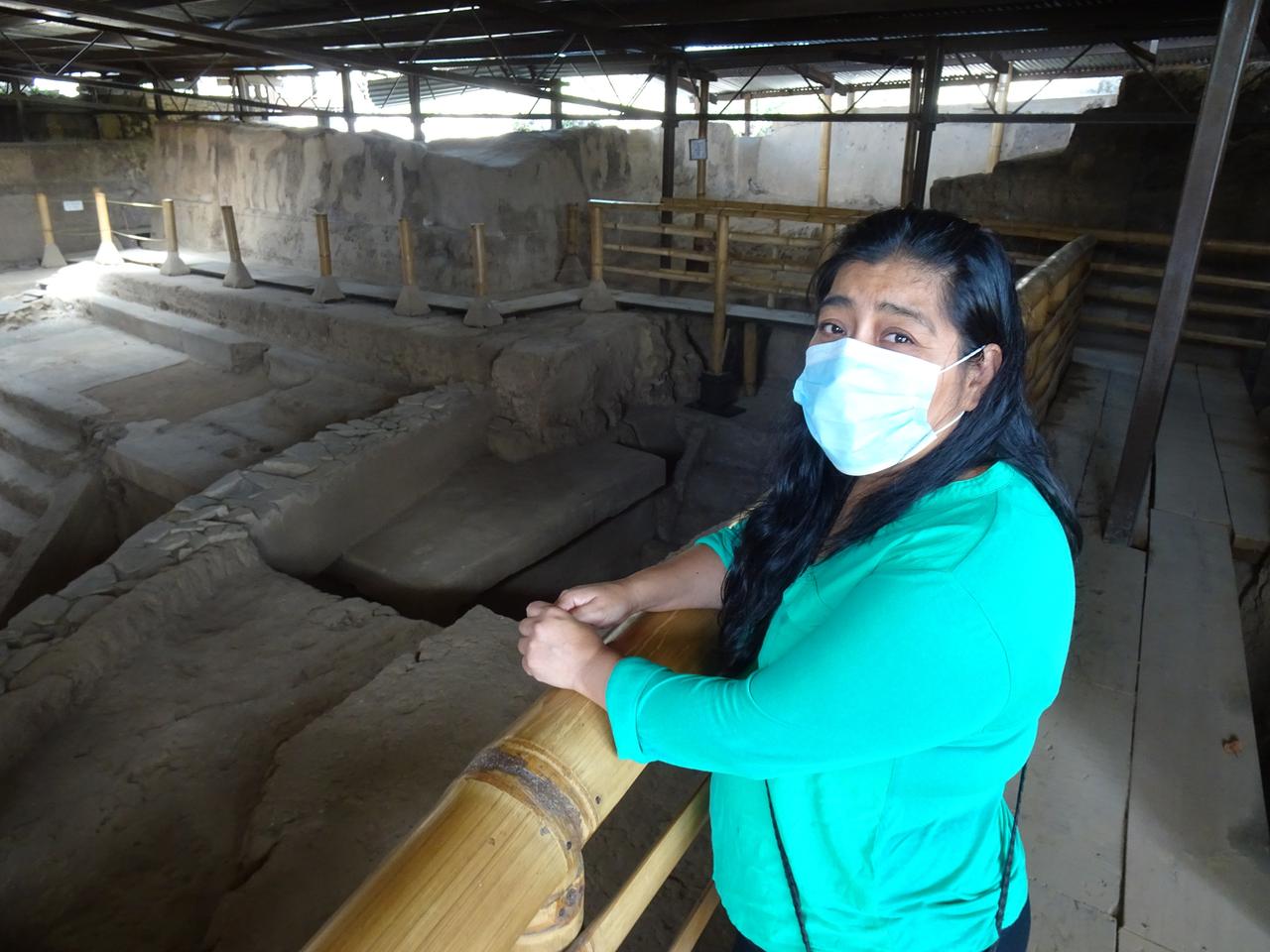 Eine Frau mit Atemschutzmaske steht an einer archäologischen Grabungsstätte vor staubigen Treppen und Kammern, die durch ein Bretterdach geschützt sind.