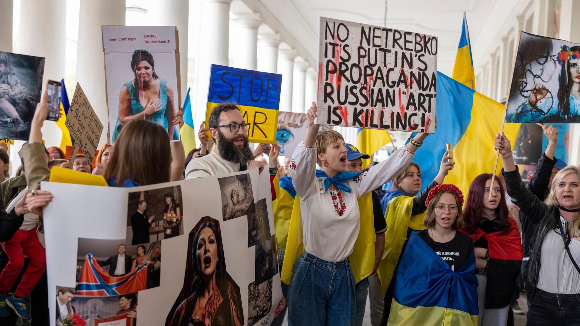 Wir sehen eine Gruppe von Demonstranten, die gegen einen Auftritt der Sopranistin Anna Netrebko protestiert. Sie sind mit Flaggen der Ukraine ausgestattet und halten Plakate gegen Netrebko in die Höhe.