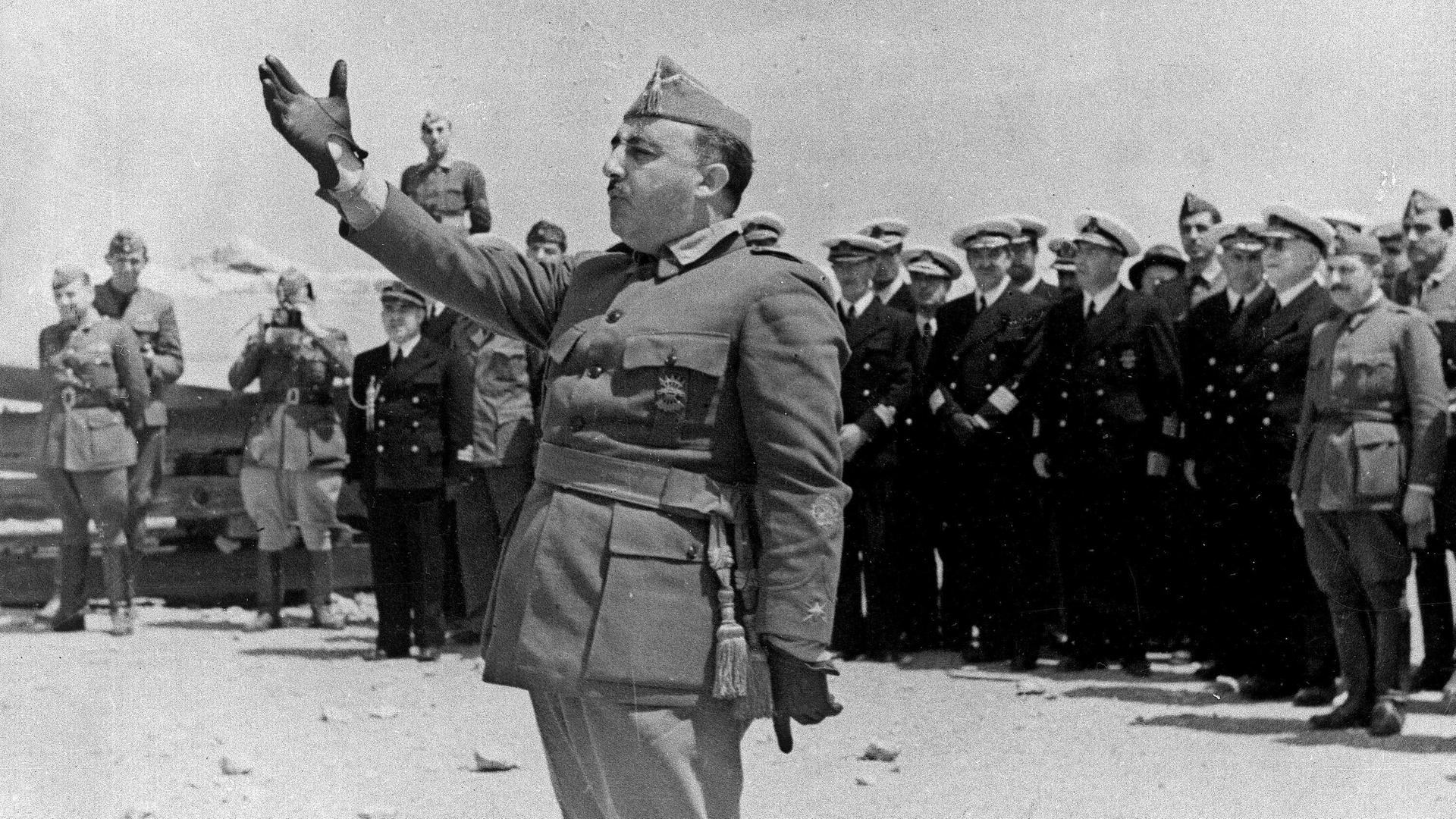 Ein Mann in militärischer Kleidung spricht, er macht eine große Geste mit seinem rechten Arm. Hinter ihm sind unscharf andere Menschen zu erkennen. Das Foto ist in schwarz weiß, aus dem Jahr 1938.  Ein Mann in militärischer Kleidung spricht, er macht eine große Geste mit seinem rechten Arm. Hinter ihm sind unscharf andere Menschen zu erkennen. Das Foto ist in schwarz weiß, aus dem Jahr 1938.