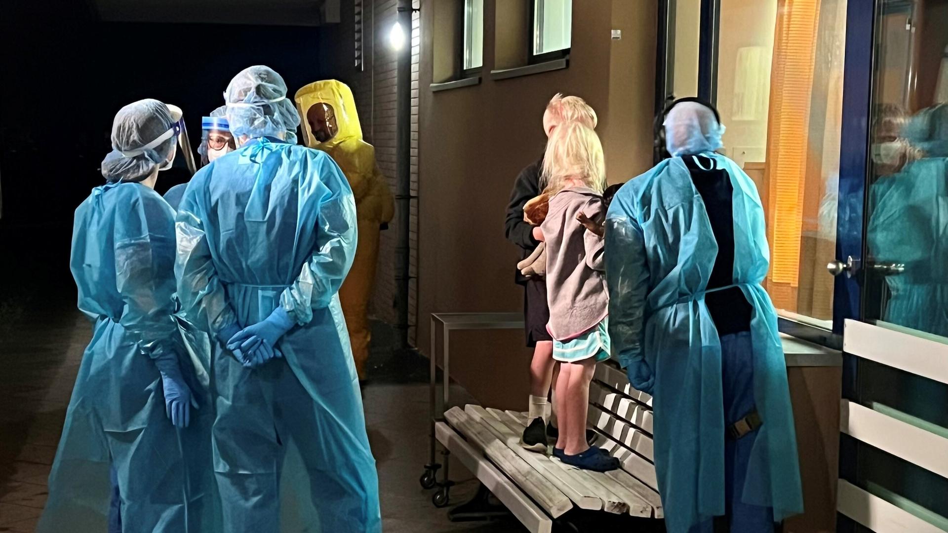 Zwei Kinder stehen auf einer Bank in einem Flur und schauen durch ein großes Fenster in ein Zimmer. Neben ihnen sind mit Kitteln, Hauben, Gesichtsschutz und Handschuhen geschützte Menschen. 