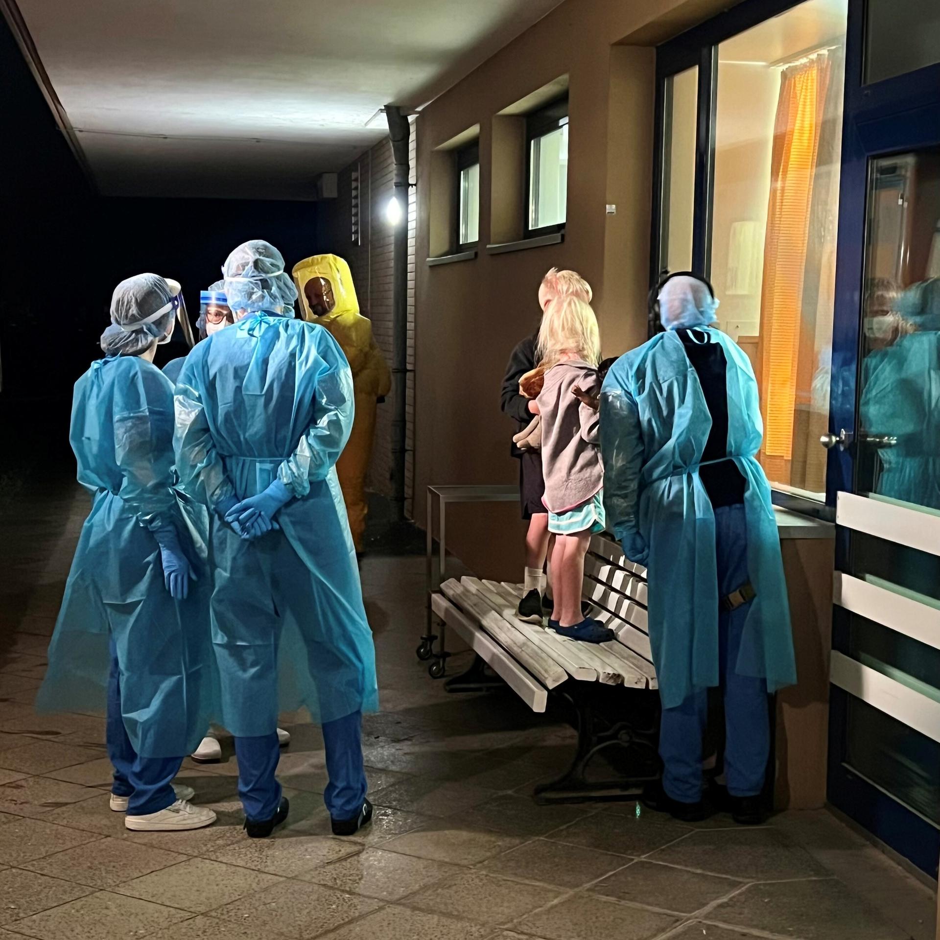Zwei Kinder stehen auf einer Bank in einem Flur und schauen durch ein großes Fenster in ein Zimmer. Neben ihnen sind mit Kitteln, Hauben, Gesichtsschutz und Handschuhen geschützte Menschen. 