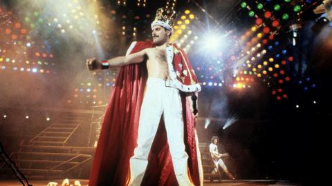 Queen-Frontmann Freddie Mercury steht in königlicher Robe und mit Krone auf dem Kopf auf der bunt beleuchteten Konzertbühne