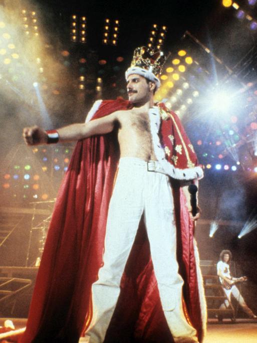 Queen-Frontmann Freddie Mercury steht in königlicher Robe und mit Krone auf dem Kopf auf der bunt beleuchteten Konzertbühne Queen-Frontmann Freddie Mercury steht in königlicher Robe und mit Krone auf dem Kopf auf der bunt beleuchteten Konzertbühne