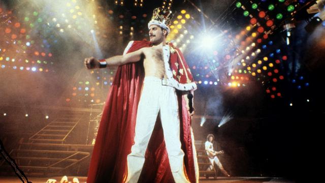 Queen-Frontmann Freddie Mercury steht in königlicher Robe und mit Krone auf dem Kopf auf der bunt beleuchteten Konzertbühne Queen-Frontmann Freddie Mercury steht in königlicher Robe und mit Krone auf dem Kopf auf der bunt beleuchteten Konzertbühne