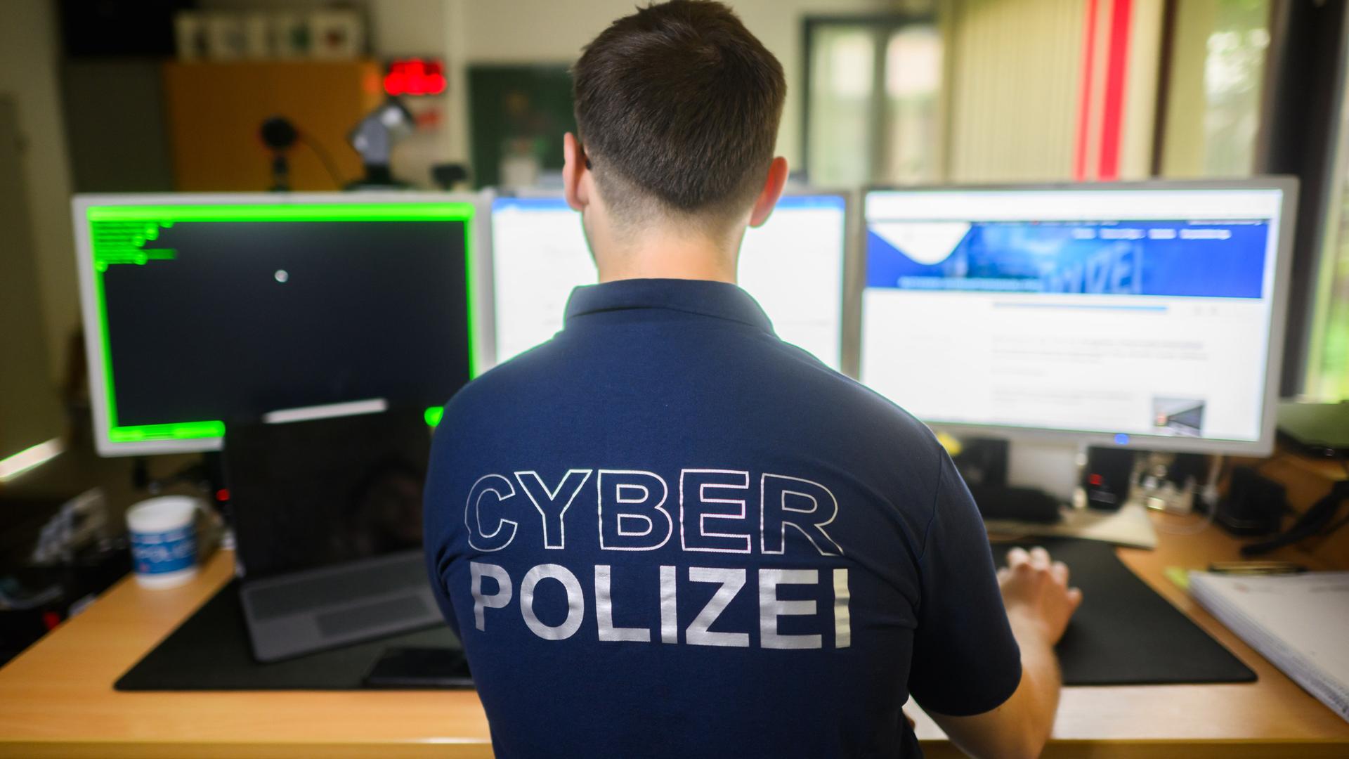 Ein Polizist steht mit einem Poloshirt mit der Aufschrift „Cyber Polizei“ an einem Computer.