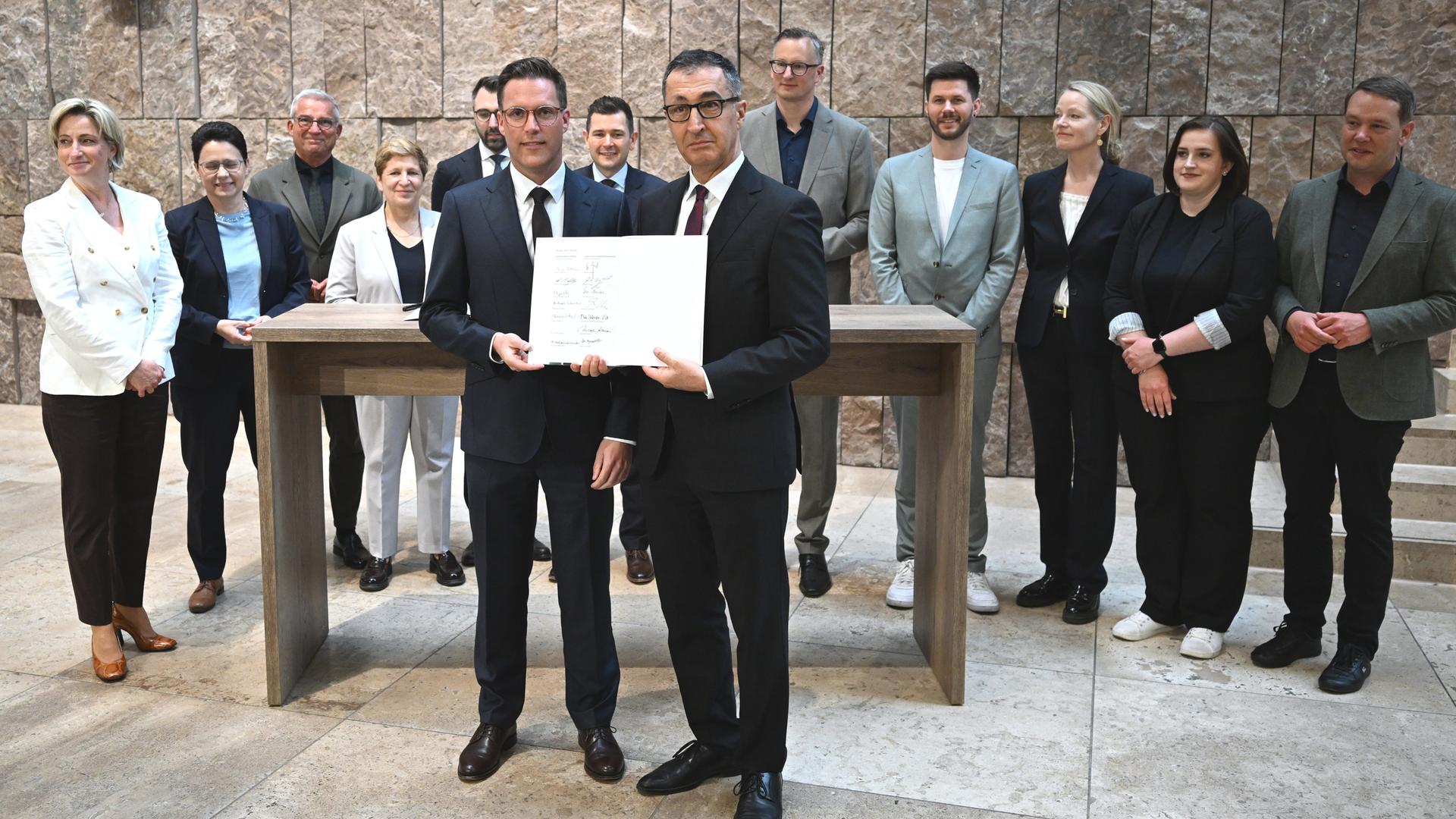 Der designierte Ministerpräsident Cem Özdemir (Grüne, r.) und sein künftiger Stellvertreter Manuel Hagel (CDU) halten den Koalitionsvertrag in die Kamera.