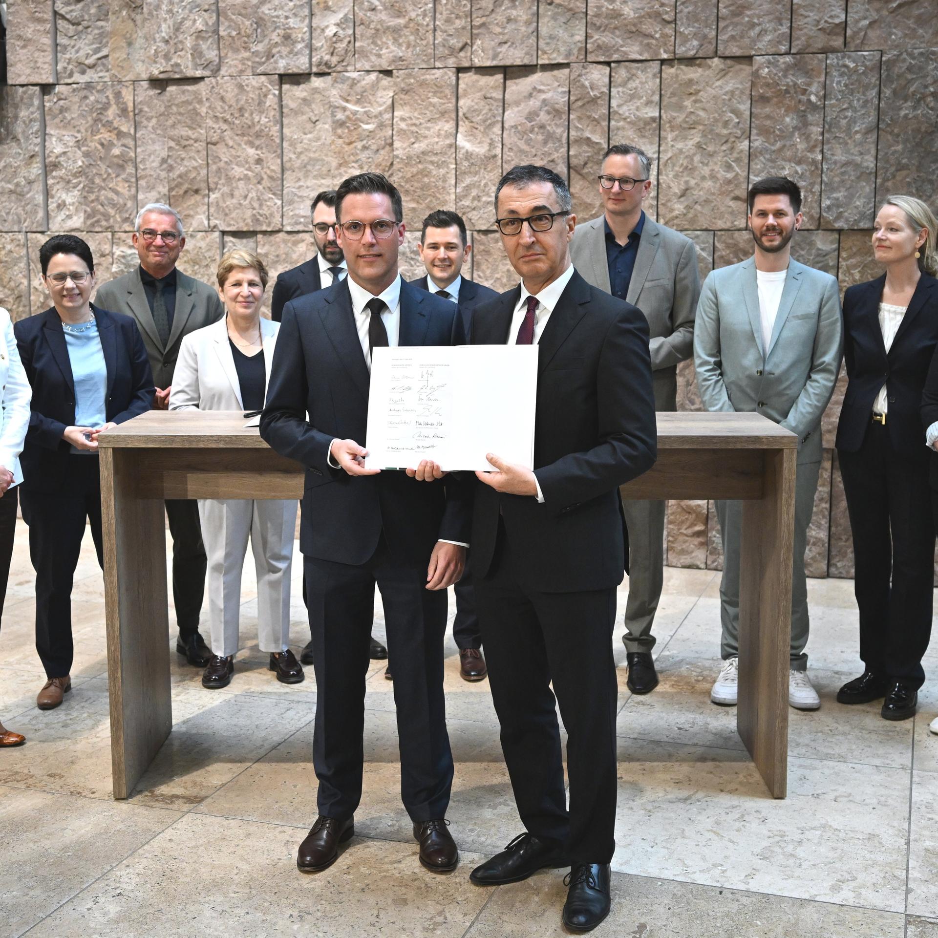 Manuel Hagel (CDU,M,l) und der designierte Ministerpräsident Cem Özdemir (Bündnis 90 / Die Grünen) halten den Koalitionsvertrag zur Bildung einer Landesregierung in Baden-Württemberg in den Händen. Hinter ihnen stehen mehrere Personen.