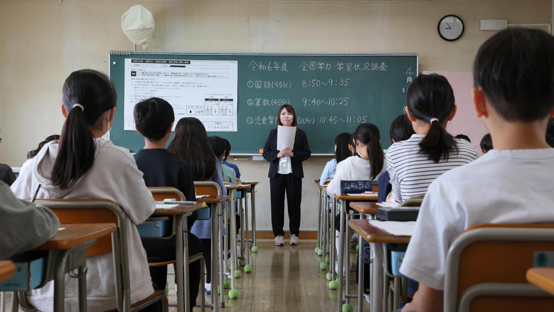 Blick in ein Klassenzimmer einer japanischen Grundschule von der hinteren Stuhlreihe aus. Vorne steht eine Lehrerin vor Aufgaben an einer Tafel Blick in ein Klassenzimmer einer japanischen Grundschule von der hinteren Stuhlreihe aus. Vorne steht eine Lehrerin vor Aufgaben an einer Tafel