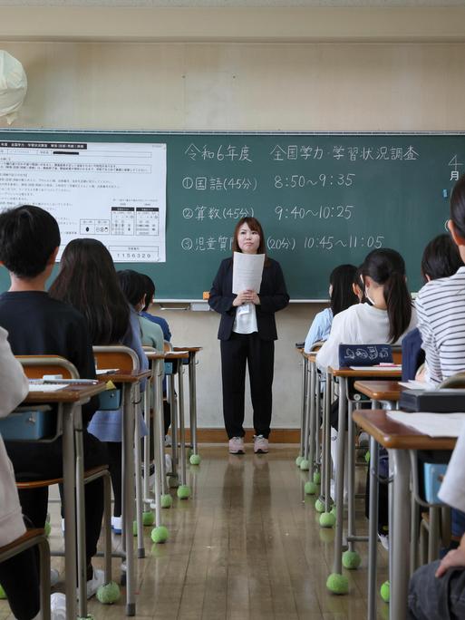 Blick in ein Klassenzimmer einer japanischen Grundschule von der hinteren Stuhlreihe aus. Vorne steht eine Lehrerin vor Aufgaben an einer Tafel
