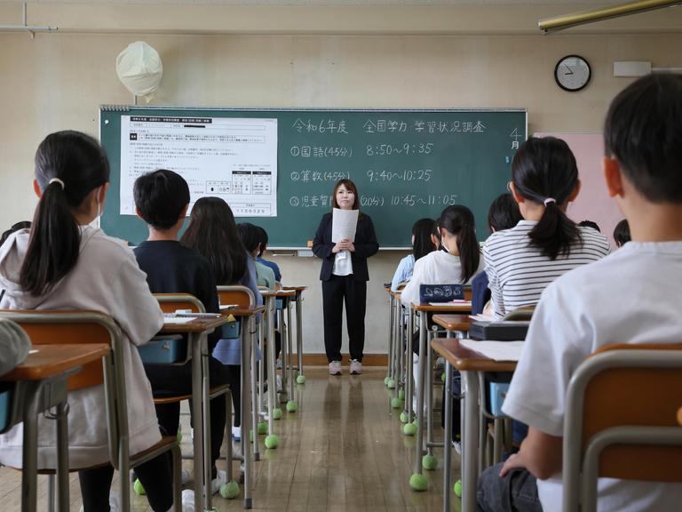 Blick in ein Klassenzimmer einer japanischen Grundschule von der hinteren Stuhlreihe aus. Vorne steht eine Lehrerin vor Aufgaben an einer Tafel