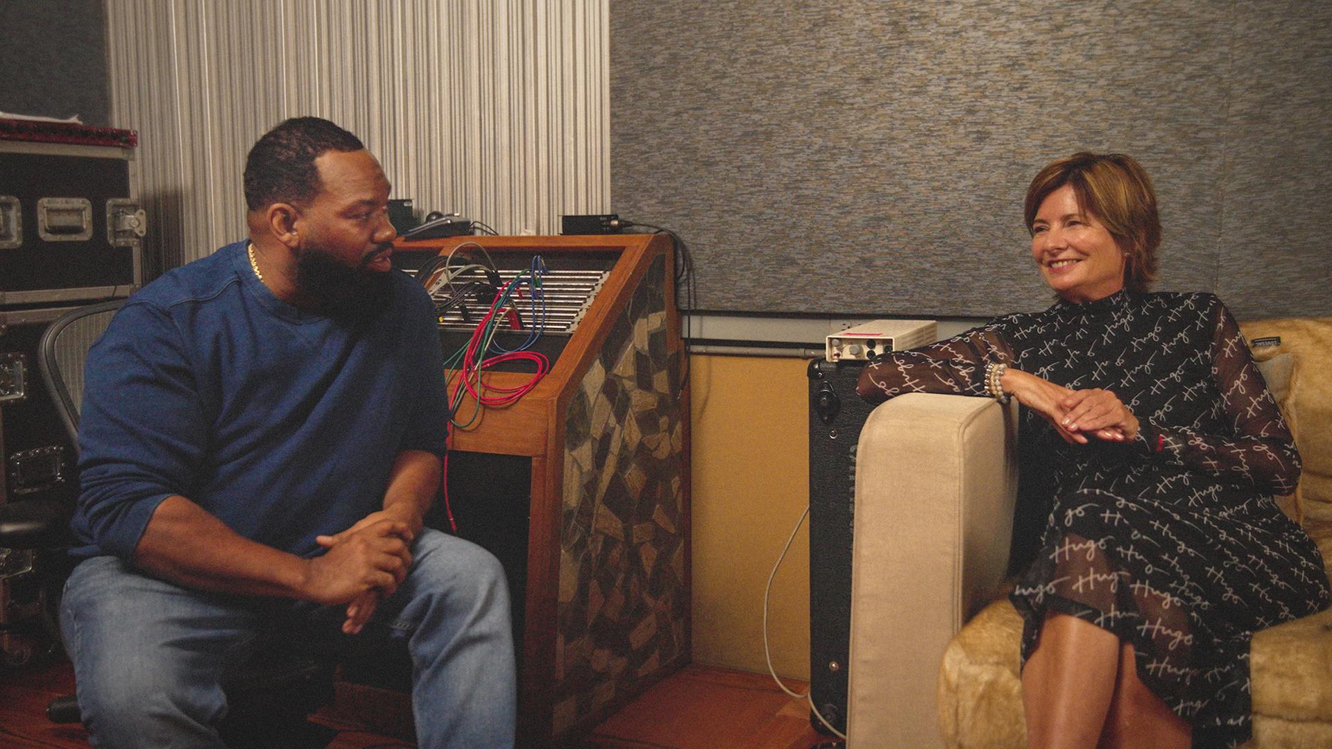 Das Foto zeigt Eva Ries im vornehmen braunen Kleid, sitzend in einem Sessel eines Tonstudios. Auf einem Stuhl links daneben sitzt Rapper Raekwon, mit blauer Jeans und blauem Pullover. 