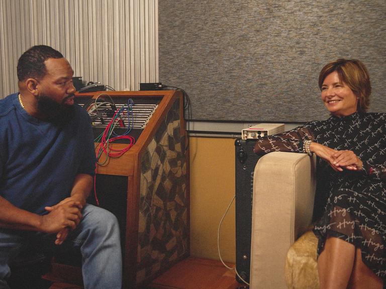 Das Foto zeigt Eva Ries im vornehmen braunen Kleid, sitzend in einem Sessel eines Tonstudios. Auf einem Stuhl links daneben sitzt Rapper Raekwon, mit blauer Jeans und blauem Pullover. 