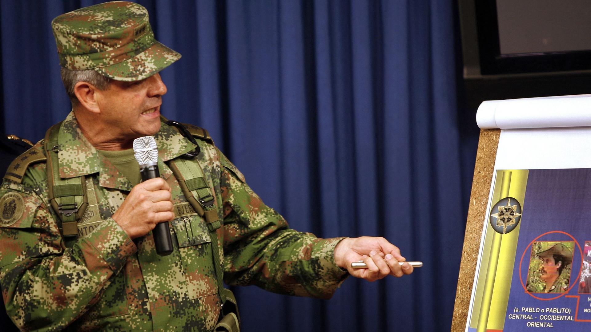 Der ehemalige kolumbianische General Mario Montoya steht vor einem Flipchart, hat ein Mikrofon in der Hand und erklärt etwas. Er trägt einen Tarnanzug.