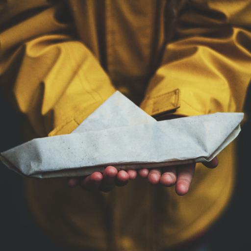 Ein Kind in einem gelben Regenmantel hält ein Papierschiff in Händen.