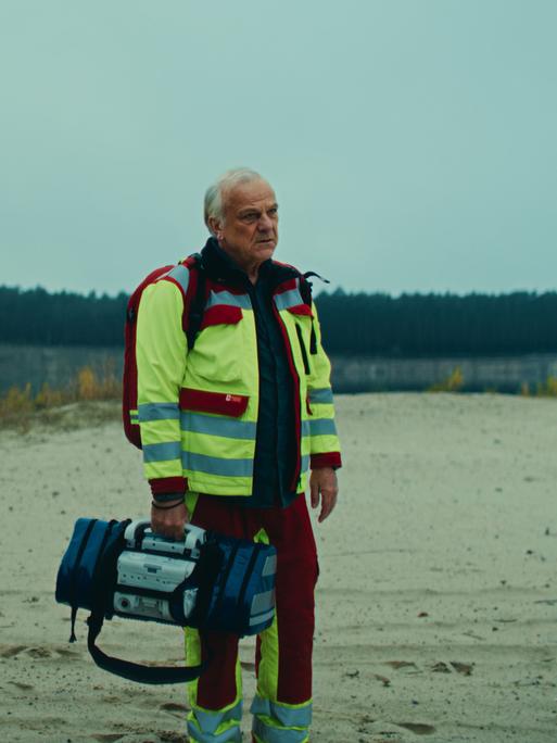 Szene aus der Serie "Krank Berlin" zeigt zwei Notfallsanitäter in einer unwirtlichen Landschaft mit sandigem Boden und einem Wäldchen im Hintergrund.