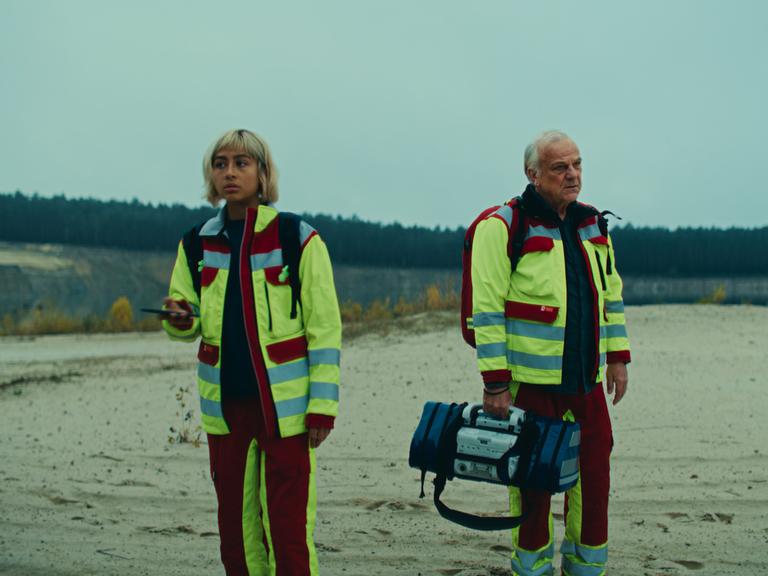 Szene aus der Serie "Krank Berlin" zeigt zwei Notfallsanitäter in einer unwirtlichen Landschaft mit sandigem Boden und einem Wäldchen im Hintergrund.