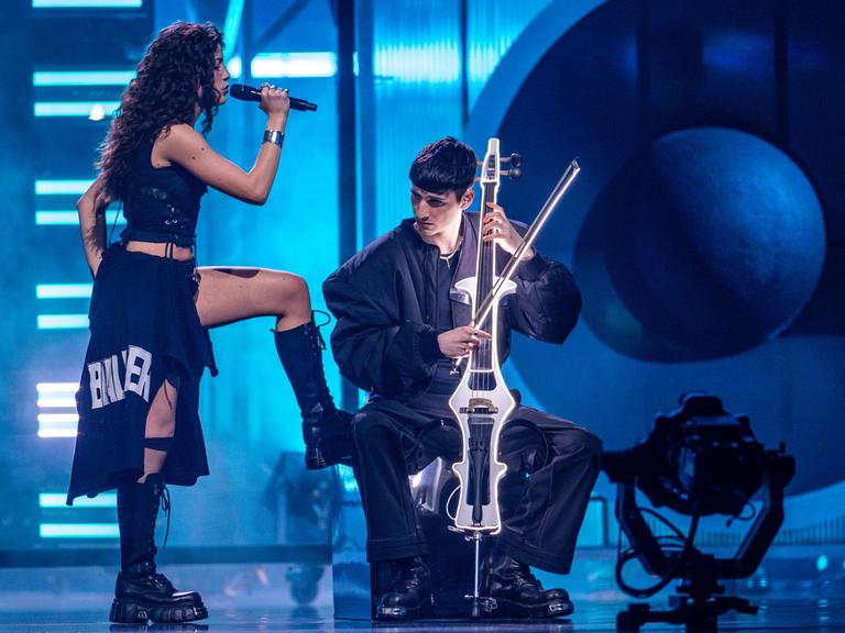 Das Duo Abor & Tynna während eines Auftritts. „Abor“ spielt Cello im Sitzen, während seine Schwester „Tynna“ stehend singt und ein Bein auf seinen Stuhl stellt. 