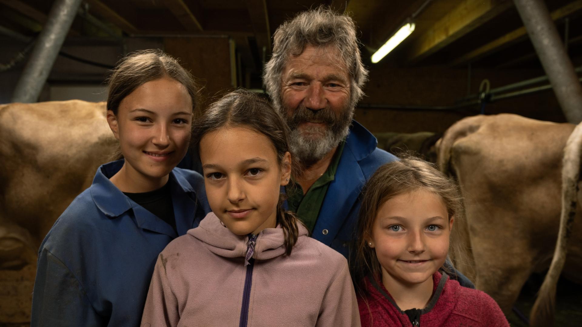 Hermann Bickel, ein älterer Herr in blauer Arbeitsjacke, und drei junge Mädchen lächeln in die Kamera. Im Hintergrund Kühe im Stall. Hermann Bickel, ein älterer Herr in blauer Arbeitsjacke, und drei junge Mädchen lächeln in die Kamera. Im Hintergrund Kühe im Stall.