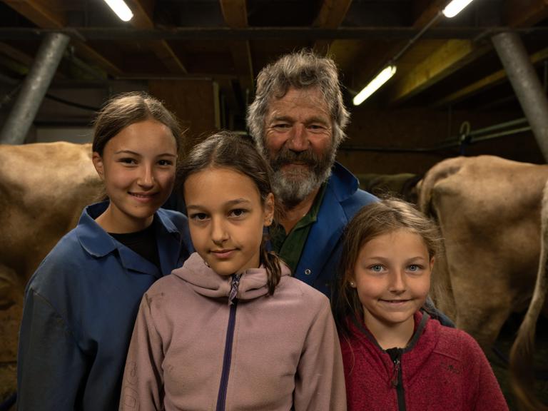 Hermann Bickel, ein älterer Herr in blauer Arbeitsjacke, und drei junge Mädchen lächeln in die Kamera. Im Hintergrund Kühe im Stall.