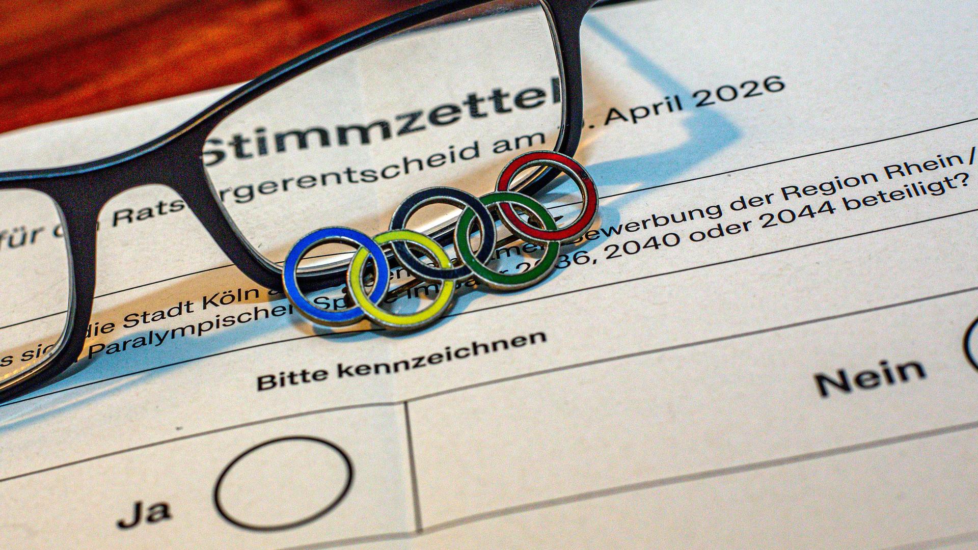 Großaufnahme eines Stimmzettels zum Referendum, ob die Region Rhein/Ruhr sich als Austragungsort für die Olympischen Spiele bewerben soll. Auf dem Zettel liegt eine Brille und ein Anstecker mit den fünf olympischen Ringen