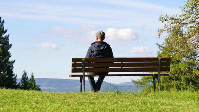 Ein älterer Herr sitzt mit dem Rücken zum Betrachter auf einer Parkbank mit Blick in die Ferne.